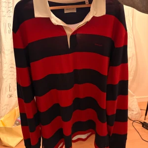 Rugby-tröja från GANT i röd/marinblå - Säljer en klassisk långärmad rugbytröja från GANT med breda röda och marinblå ränder, vit krage och öppen halsringning. Tröjan har en relaxed fit i storlek 2XL och är perfekt för dig som gillar preppy stil. Materialet är mjukt och skönt, i bomull. 