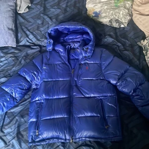 Blå pufferjacka från Polo Ralph Lauren - Säljer en blå pufferjacka från Polo Ralph Lauren med huva och dragkedja. Jackan är glansig med logga på bröstet. Mysigaste jackan jag haft kostar ungefär 6000 ny, använt i en månad ungefär! Kom dm vid minsta lilla intresse!