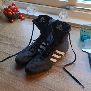 Adidas boxningsskor svarta strl 38 - Säljer ett par svarta Adidas boxningsskor med vita ränder och hög skaft. Skorna har snörning hela vägen upp och är tillverkade i lätt syntetmaterial med meshpaneler för bra ventilation. Perfekta för dig som tränar boxning och vill ha grymt grepp och smidighet. På bilderna är den dammig då dom inte används på typ 2år men har använt dom ungefär 2-3 gånger