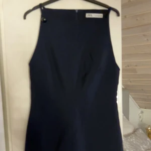Mörkblå klänning från Zara - En stilren mörkblå klänning från Zara med minimalistisk design och hög halsringning. Klänningen har en knappdetalj vid axeln och är ärmlös, vilket ger en clean och modern look. Perfekt för dig som gillar enkelhet och klassiska snitt.