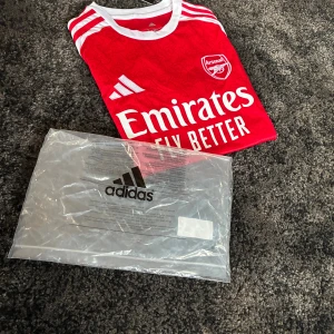 Arsenal hemmatröja Adidas S - •	Storlek: S 	•	Material: 100% polyester (AEROREADY – fukttransporterande) 	•	Nyskick, oanvänd med taggar kvar 	•	Levereras med originalförpackning från Adidas 	•	Färger: Röd med vita ärmar, klassisk Arsenal-design 	•	Sponsorer: Emirates Fly Better på framsidan och Visit Rwanda på ärmen  Perfekt både att bära på matchdag eller som samlartröja!  📦 Skickas snabbt och välpackat. 