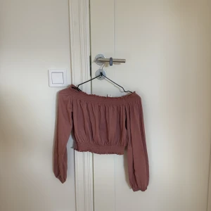Rosa offshoulder croppad blus - Säljer en söt croppad blus i dammig rosa färg med offshoulder och långa ärmar. Blusen har smock vid halsringning och midja samt små volangdetaljer upptill och nedtill. 💘
