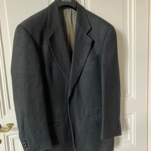 Mörkgrå kavaj från Hugo Boss - Snygg mörkgrå kavaj från Hugo Boss i modellen Maloma. Kavajen har klassisk passform med tre knappar vid ärmslut, bröstficka och två fickor framtill. Tillverkad i en blandning av linne och ull för en stilren och exklusiv känsla. Storleken är snarare 48-50 än 46. 