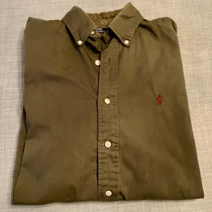 Olivgrön skjorta från Polo Ralph Lauren - Olivgrön skjorta från Polo Ralph Lauren med klassisk krage och vita knappar framtill. Broderad röd logga på bröstet ger en snygg detalj. Skjortan är långärmad och oversized, snarare Large än Medium. Se mått på bild.