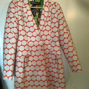Desigual kavaj med stora prickar 38 size - Fet kavaj från Desigual med stora vita prickar på orange botten och två knappar framtill. Insidan har ett färgstarkt, mönstrat foder med blommor och fjärilar. Jackan har klassisk krage och långa ärmar. Materialmix av bomull och polyester.