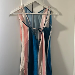 Randig klänning med snörning - Snygg långklänning med breda vertikala ränder i blått, rosa och vitt. Klänningen har tunna axelband, väljbar djup v-ringning och snörning framtill baktill. Lätt och flowy känsla, där du ändrar längden på banden i knytet vid ryggen