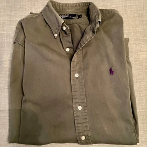 Olivgrön skjorta från Polo Ralph Lauren - Olivgrön skjorta från Polo Ralph Lauren med klassisk krage och lila broderad logga på bröstet. Skjortan har långa ärmar, vita knappar framtill och är tillverkad i mjuk bomull.  Skjortan är långärmad och oversized, snarare Large än Medium. Se mått på bild. Obs, bilderna med mått visar tvillingskjorta i annan färg. 