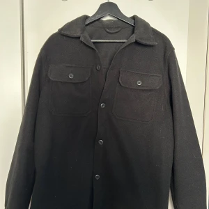 Svart overshirt  - Stilren svart overshirt med klassisk krage, två bröstfickor med knappar och hel knappslå framtill. Tillverkad i ett mjukt ullmaterial. Perfekt för lager-på-lager och enkel att styla.