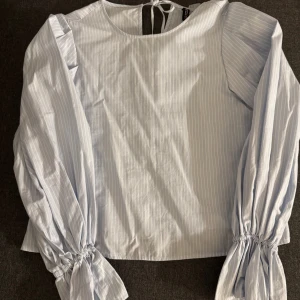 Randig blus med puffärm från H&M Divided - Säljer en ljusblå och vitrandig blus från H&M Divided i storlek XS. Blusen har långa puffärmar med knytband i ryggen. Tillverkad i bomull, perfekt för en trendig och fräsch look. Hel ny tröja har aldrig använt.