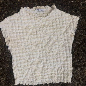 Vit strukturerad topp från Zara - Säljer en vit topp från Zara med unik bubblig struktur och lätt vågiga kanter. Toppen är ärmlös och har en högre halsringning. Perfekt för dig som gillar en stilren men ändå intressant look.
