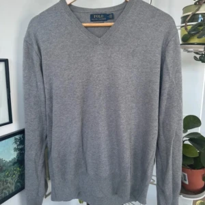 Grå tröja Polo Ralph Lauren - Klassisk grå v-ringad tröja från Polo Ralph Lauren i 100% pima bomull. Tröjan har diskret broderad logga på bröstet och ribbade muddar vid ärmslut och nederkant. Perfekt för lager-på-lager och enkel att matcha med allt.