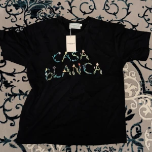 Svart t-shirt från Casablanca - Svart t-shirt från Casablanca med färgglad broderad text 'CASA BLANCA' på bröstet, där bokstäverna är dekorerade med blommor och svampar. Klassisk passform och rund hals. Perfekt för dig som vill sticka ut med en cool detalj.