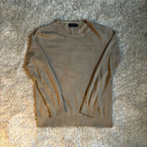 Beige stickad tröja från Riley - En stilren beige stickad tröja från Riley med rund halsringning och långa ärmar. Tröjan har ribbade muddar vid ärmslut och nederkant, perfekt för lager-på-lager stil. 