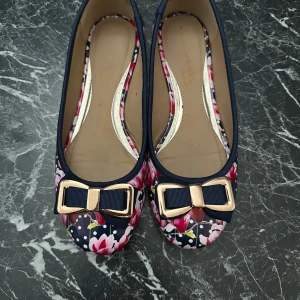 Blommiga ballerinaskor med rosett - Snygga ballerinaskor med marinblå och rosa blommigt mönster, dekorerade med en stor marinblå rosett med guldfärgad detalj framtill. Skorna har rund tå och platt sula.Bekväma och lätta att bära. Köptes på magazine i Köpenhamn.
