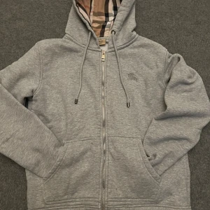 Grå hoodie från Burberry med dragkedja - Säljer en grå hoodie från Burberry med klassiskt rutigt foder i huvan. Tröjan har dragkedja framtill, snörning i huvan och två fickor. Perfekt för dig som gillar stilrena och exklusiva plagg med en twist.