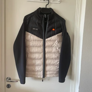 Svart och beige pufferjacka från Ellesse - Snygg pufferjacka från Ellesse i svart och beige med quiltad front och ärmar i svart. Jackan har dragkedja hela vägen, hög krage och Ellesse-logga på bröstet. Tillverkad i 100% polyester, perfekt för kyligare dagar.