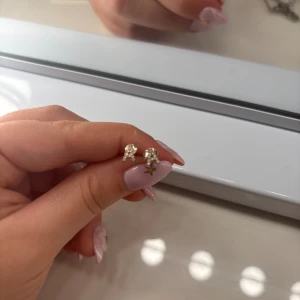 Söta silvriga örhängen med ett A - Ett par små silvriga örhängen i form av söta figurer, där ena örhänget har en liten stjärna hängande. De är glittriga och perfekta för att ge din look lite extra shine. Passar dig som gillar detaljer och unika smycken.