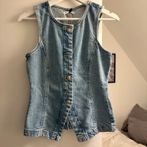 Jeans topp - Snygg ljusblå jeansväst i klassisk denim med markerade sömmar och silverfärgade knappar framtill. Västen är ärmlös och har en figurnära passform med lätt v-ringad hals. Perfekt att styla över en t-shirt eller klänning.