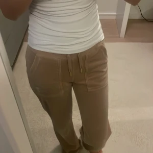 Beiga mjukisbyxor Juicy Couture XXS - Beiga mjukisbyxor från Juicy Couture i supermjuk velour med snörning i midjan och guldiga detaljer på bakfickorna. Byxorna har raka ben och stora fickor framtill, perfekt för en chill och trendig look.