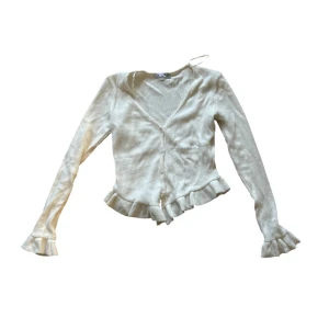 Beige kofta med volang - ZARA - Supersöt beige kofta från Zara med v-ringning, knappar framtill och volangdetaljer vid nederkant och ärmslut. Modellen är slim och har långa ärmar. Perfekt att styla med jeans för en chill look.