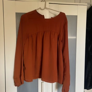 Rostbrun blus från Angelology S - Säljer en snygg rostbrun blus från Angelology i storlek S. Blusen har rund hals, långa ärmar och ett lätt rynkat parti under bysten som ger en chill och flowy vibe. Materialet är mjukt och har ett lätt glansigt utseende, perfekt för dig som gillar stilrena plagg med lite extra touch.