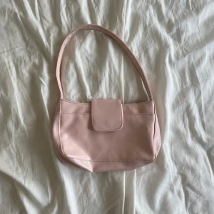 Ljusrosa axelväska i syntet - Söt ljusrosa axelväska med enkel design och mjuka former. Väskan har ett brett axelband och stängs med ett lock framtill. Perfekt storlek för det viktigaste och ger en clean look till din outfit.