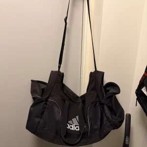 Svart träningsväska från Adidas - Stor och rymlig svart träningsväska från Adidas med klassisk logga framtill. Väskan har flera fack, justerbar axelrem och detaljer i syntetmaterial. Perfekt för gymmet eller sportaktiviteter, med smarta förvaringsmöjligheter och stilren design.