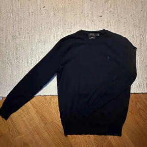 Svart merinoullströja från Polo Ralph Lauren - Slim fit svart tröja från Polo Ralph Lauren i tvättbar merinoull. Klassisk rund halsringning och diskret broderad logga på bröstet. Långärmad modell med ribbade muddar och nederkant. Perfekt för lager-på-lager under kyliga dagar.