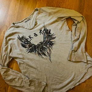 Beige långärmad tröja Zadig & Voltaire - Säljer en beige långärmad tröja från Zadig & Voltaire med coolt tryck av vingar och texten 'Rock & Roll' på bröstet. Tröjan har v-ringad hals och är tillverkad i mjuk bomull, perfekt för dig som gillar edgy stil.