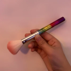 Regnbågsfärgad sminkborste - Superfin sminkborste med regnbågsfärgat handtag i metallic och mjuka syntetborst i vitt och rosa. Perfekt för att applicera puder, bronzer eller rouge. Handtaget har en snygg övergång mellan lila, rött, gult och grönt. Borsten är fluffig och lätt att jobba med.