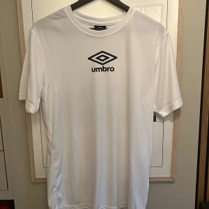 Vit Umbro t-shirt i polyester S - Snygg vit t-shirt från Umbro i storlek S. T-shirten har klassisk passform, korta ärmar och är gjord i ett lätt och sportigt polyestermaterial. Stor svart Umbro-logga framtill och en mindre logga i nacken. Perfekt för dig som gillar sportig stil.