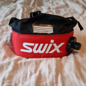 Röd och svart Swix vätskebälte - Sportig midjeväska från Swix i rött och svart med stor vit logga. Väskan har justerbart midjeband, dragkedja upptill och en stor skruvkork för vätskebehållare. Perfekt för längdskidåkning eller träning utomhus. Användes vid träning inför tjejvasan och under loppet så kan inte påstå att den är välanvänd..