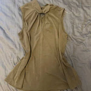 Grön ärmlös blus från H&M - Säljer en grön ärmlös blus från H&M med snygg knytdetalj vid halsen och tre klädda knappar i nacken. Blusen har en lätt utsvängd passform och är tillverkad i ett mjukt, tunt tyg som känns skönt mot huden.