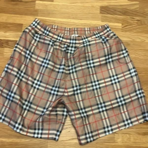 Rutig Burberry shorts i bomull - Säljer ett par klassiska Burberry shorts i storlek M med det ikoniska rutiga mönstret i beige, svart, vit och röd. De har elastisk midja och är tillverkade i mjuk bomull med meshfoder. Perfekta för dig som vill ha en clean och lyxig vibe.