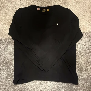 Polo Ralph Lauren Tröja Svart - Svart långärmad tröja från Polo Ralph Lauren med klassisk vit broderad logga på bröstet. Tröjan har rund halsringning och är gjord i mjuk bomull, perfekt för en clean och stilren look.