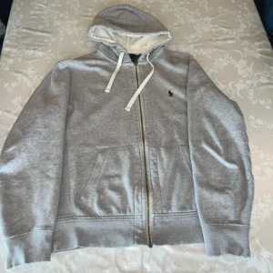 Grå hoodie från Polo Ralph Lauren M - Klassisk grå hoodie från Polo Ralph Lauren med dragkedja, vit snörning i huvan och broderad logga på bröstet. Mjuk insida och känguruficka framtill. Tillverkad i bomull och polyester, perfekt för chill dagar. Lite trasig vid sömnen vid armen ser på sista bilderna 