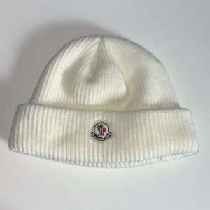 Vit Moncler mössa - Snygg vit mössa från Moncler med ribbad struktur och uppvikt kant. Framtill sitter Monclers klassiska broderade logotyp i blått och rött. Mössan är tillverkad i mjuk ull och har en clean, stilren look som passar perfekt till kalla dagar. Pris kan diskuteras!