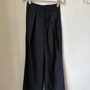 Svarta wide kostymbyxor från Lemaire - Snygga svarta kostymbyxor från Lemaire i storlek 34. Byxorna har en wide passform med hög midja och pressveck framtill. Materialet är en lyxig mix av ull, spandex och viskos som ger både komfort och stil. Perfekta för dig som gillar minimalistisk och clean look. Never used. 