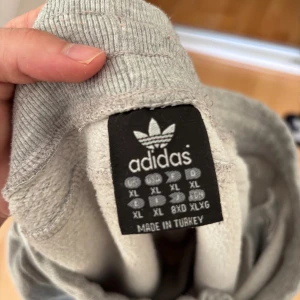 Grå Adidas mjukisbyxor  - FÖR DIG SOM VILL ANVÄNDA DE FÖR ARBETE ELLER ÄR KREATIV O KAN FÄRGA DEM FINT MED KLÄDFÄRG. Grå mjukisbyxor från Adidas i storlek XL med jag valde M då den ABSOLUT är mycket mindre. Den har klassiska vita ränder längs benen, snörning i midjan och dragkedjeförsedda fickor. 