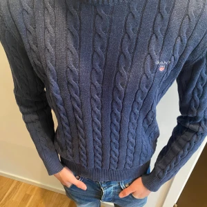 Marinblå kabelstickad tröja från Gant - Säljer en marinblå kabelstickad tröja från Gant med rund halsringning och broderad logga på bröstet. Tröjan är långärmad och har ribbade muddar vid ärmslut och nederkant. Perfekt för dig som gillar klassisk och stilren look.
