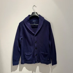 Marinblå kofta från Gant, storlek M - Snygg marinblå kofta från Gant med klassisk krage och tre knappar framtill. Koftan har två fickor och är tillverkad i mjuk bomull. Perfekt för lager-på-lager och passar till många olika stilar. Hör av er vid frågor