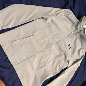Under Armour Jacka - Snygg ljusgrå jacka från Under Armour med huva och dragkedja hela vägen. Jackan har två sidofickor och logga på bröstet samt tryckt Under Armour-text på ryggen. Perfekt för träning eller när du vill ha en sportig look. Säljs då den inte passar mig.