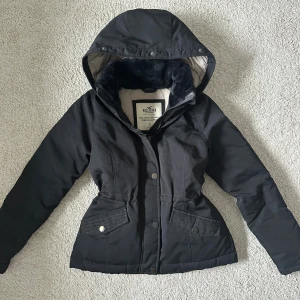Svart dunjacka från Hollister med huva, S - Svart dunjacka från Hollister med fluffigt foder i kragen och stor huva. Jackan är sparsamt använd och i mycket fint skick. Den har en snygg passform med dragsko i midjan. Garderobsrensar så kolla gärna in andra annonser!