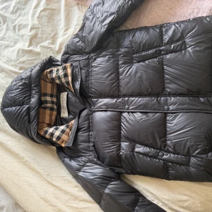 Burberry vinterjacka - Svart pufferjacka från Burberry med klassiskt rutigt foder i beige och svart. Den är helt oanvänd  och är i stl 52.