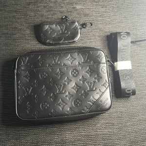 Svart axelväska skinn - Snygg svart axelväska  i präglat skinn med klassiskt monogrammönster. Väskan har en bred axelrem med logga, dragkedja och tillhörande liten plånbok. Perfekt för dig som vill ha en stilren och lyxig accessoar.