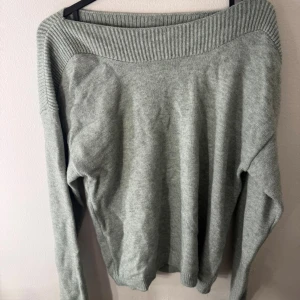 Grå stickad tröja med ribbad kant - Mysig grå stickad tröja med bred ribbad kant runt halsen och axlarna. Tröjan har långärmade ärmar och en avslappnad passform som gör den enkel att styla. Perfekt för kyliga dagar och passar till jeans eller kjol.