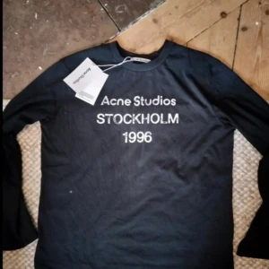 Svart långärmad tröja Acne Studios - Svart långärmad tröja från Acne Studios med vit text 'STOCKHOLM 1996' på bröstet. Enkel och stilren design, perfekt för dig som gillar klassiska streetwear-plagg med tydlig logga. Rund hals och mjukt bomullsmaterial. S men passar M också stor i storlek 