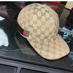 Gucci beige keps med GG-mönster - Snygg beige keps från Gucci med klassiskt GG-monogram över hela kepsen. Har gröna och röda ränder på sidan och justerbart läderband baktill. Materialet är bomull och kepsen har en rundad form med böjd skärm. Perfekt för dig som gillar lyxiga accessoarer.