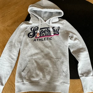 Grå Superdry hoodie med tryck - Snygg ljusgrå hoodie från Superdry med stort tryck på bröstet och japanska tecken. Tröjan har huva med snörning, magficka och ribbade muddar. Passformen är slim och materialet är mjuk bomull. Perfekt för dig som gillar sportig och clean stil.