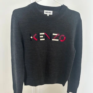 Mörkgrå ulltröja från Kenzo - Säljer en mörkgrå stickad tröja från Kenzo i 100% ull. Tröjan har rund halsringning och färgglatt broderat Kenzo-logo i rött, vitt och mörkblått framtill. Orginalpris 2900kr, aldrig använt